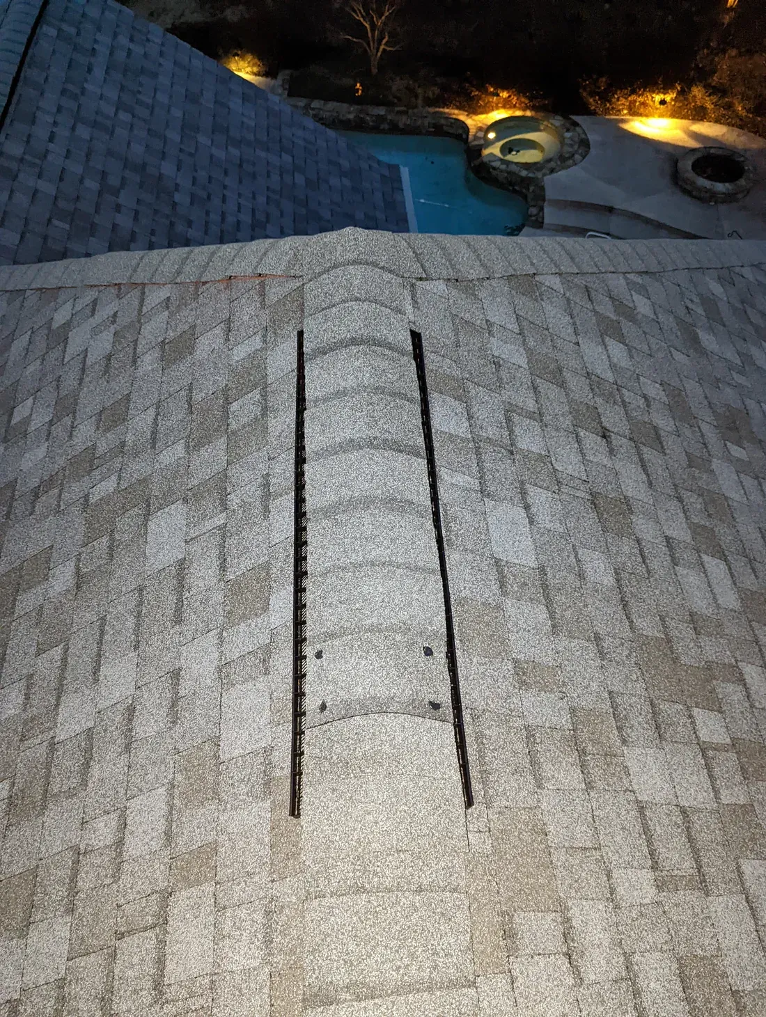 View of a light-colored shingled roof with a dark, open vent leading to a pool area at night.