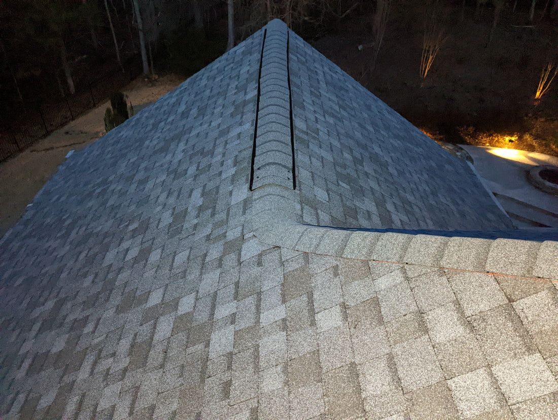 Overhead view of a roof with gray shingles. A dark ridge line runs along the top, set against a dark wooded backdrop.