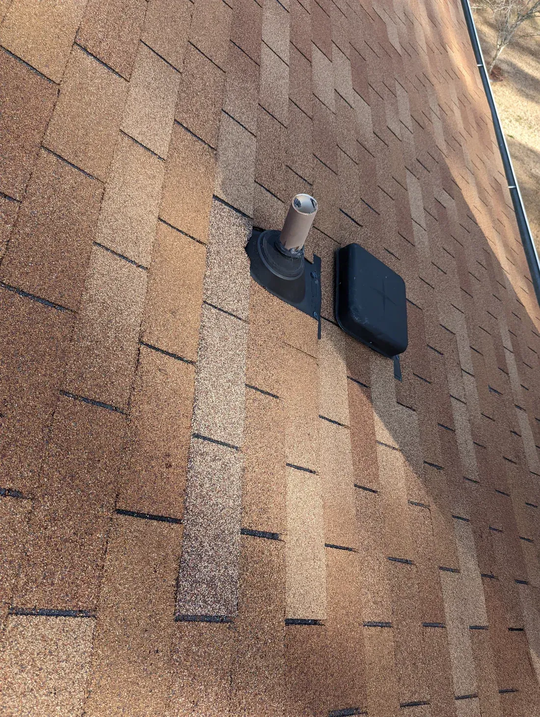 Tan asphalt shingle roof with a vent pipe and black rectangular vent.