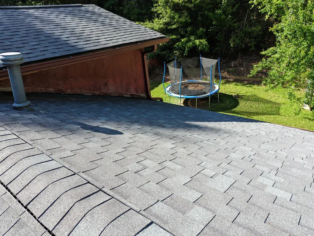 Gray asphalt shingle roof with a brown building, a trampoline, and trees in the background.
