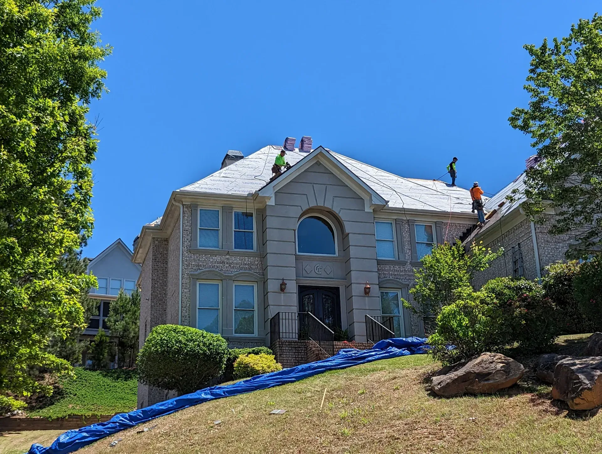 Workers replacing roof shingles on a two-story brick house under a blue sky; blue tarp on the lawn.