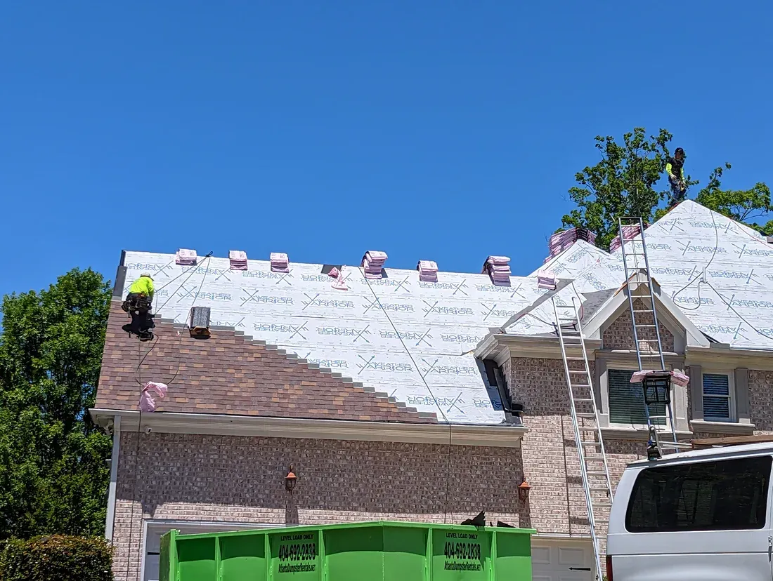 Roofing work on a house; worker on roof, ladder, green dumpster, blue sky.