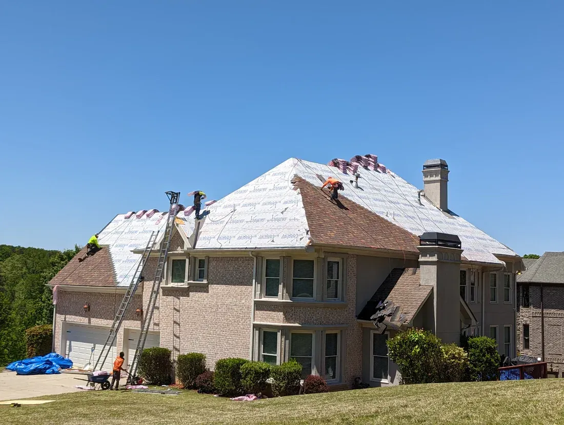 Roofers working on a two-story home, replacing shingles under a clear, blue sky.