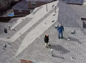Two people stand on a gray shingle roof. Other buildings are in the background.