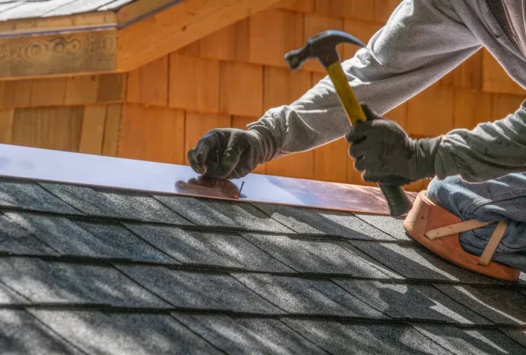 Roofer hammering copper flashing onto a dark shingled roof, outdoors.