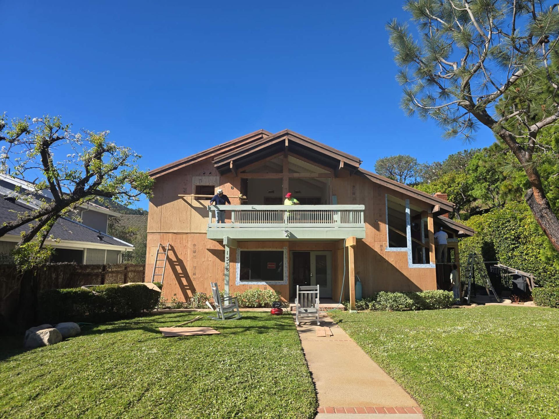 Two people work on the balcony of a two-story tan house with a tiled roof, set against a backdrop of trees and a lawn.