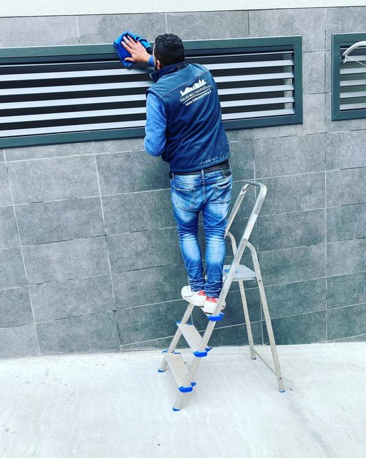 Uomo su una scala a pioli che pulisce le lamelle di ventilazione esterne con un panno blu.
