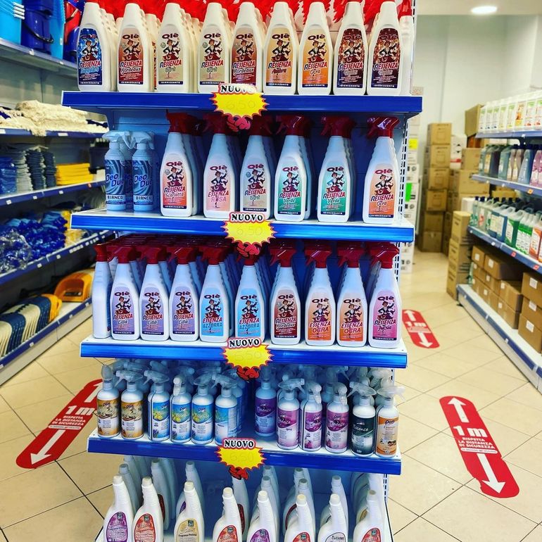 Scaffali pieni di colorati flaconi di detergente spray in un negozio, con cartelli di vendita e indicatori di distanziamento sociale sul pavimento.