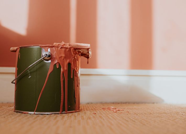 Un barattolo di vernice verde con vernice color corallo che gocciola, appoggiato su un tappeto vicino a una parete color corallo appena dipinta.