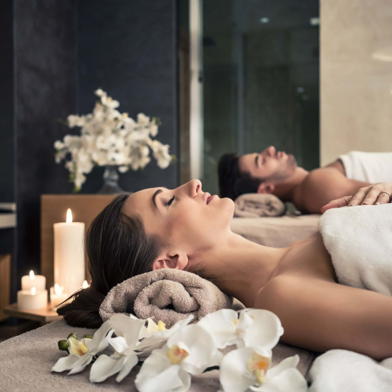 A couple relaxing at a spa. A woman and a man are lying on massage tables, eyes closed, near candles and flowers.