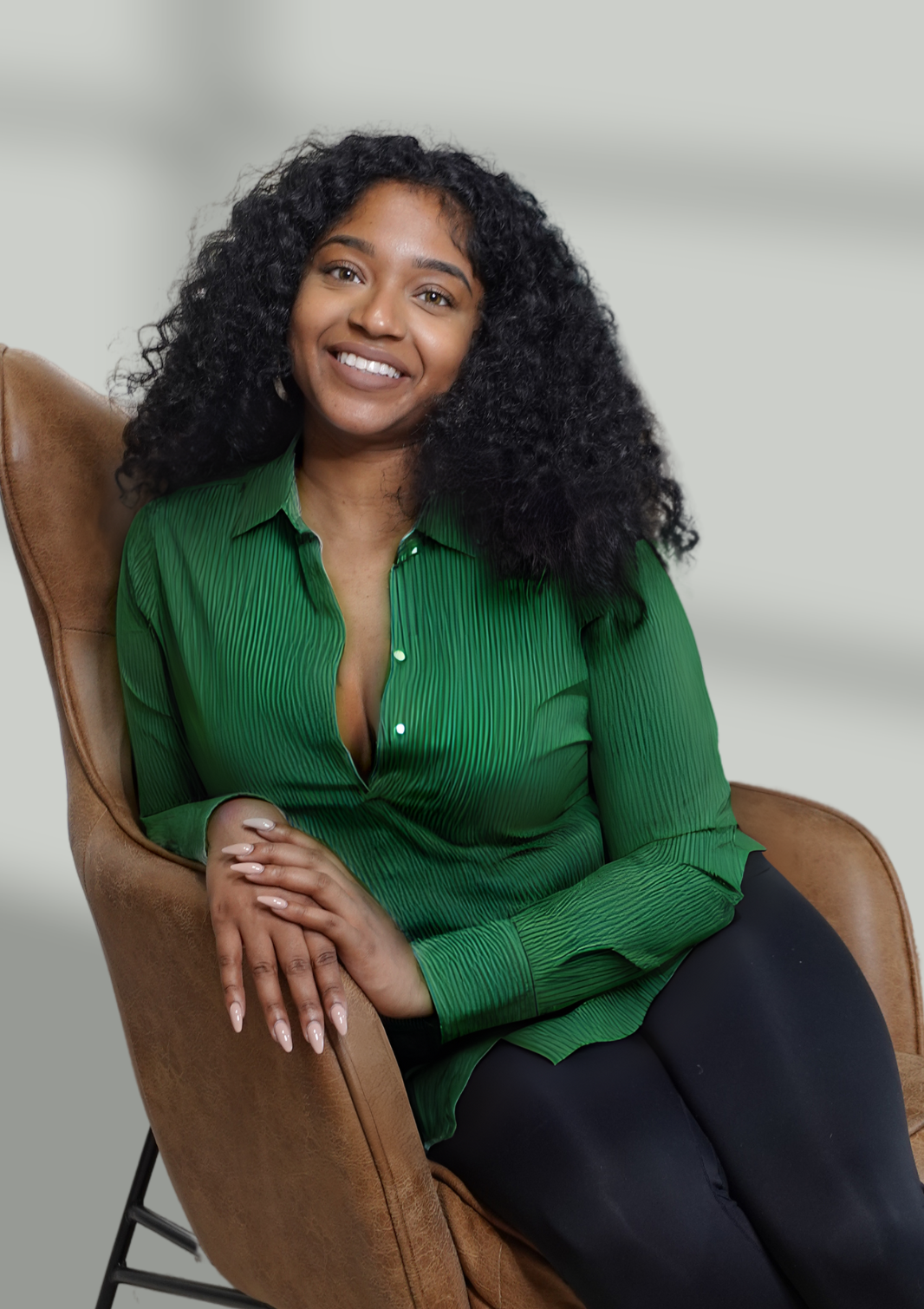 Jess sits in a brown leather chair against a soft gray background. She has curly black hair and is smiling while wearing a green blouse.