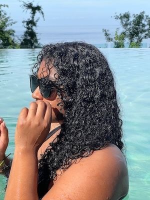 Jess wears black sunglasses in a pale blue infinity pool with wet curly hair and trees near the water in the background.