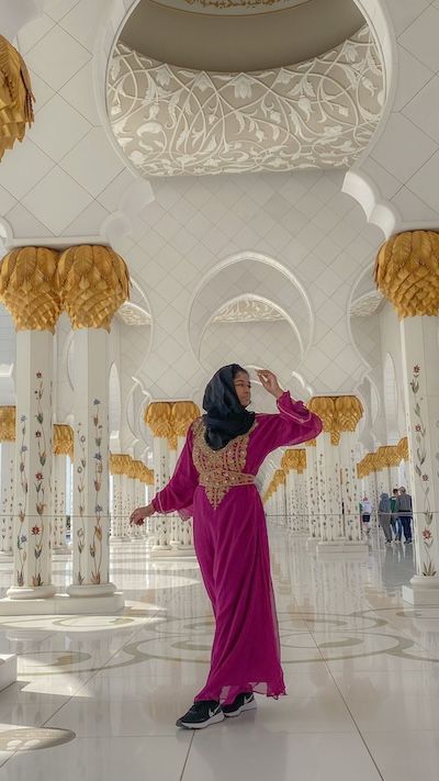 Jess poses in a pink dress and black headscarf inside a white mosque with floral columns, arches, and gold chandeliers.
