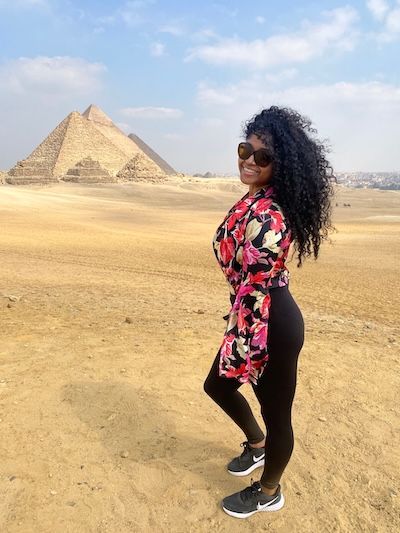 Jess smiles in front of the Pyramids of Giza while wearing sunglasses, a floral top, black leggings, and sneakers.