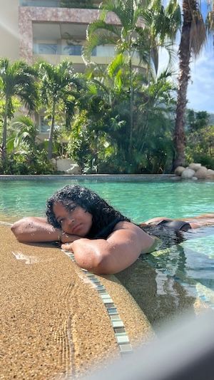 Jess rests at the edge of a resort pool in a black swimsuit with palm trees and a hotel building behind her.