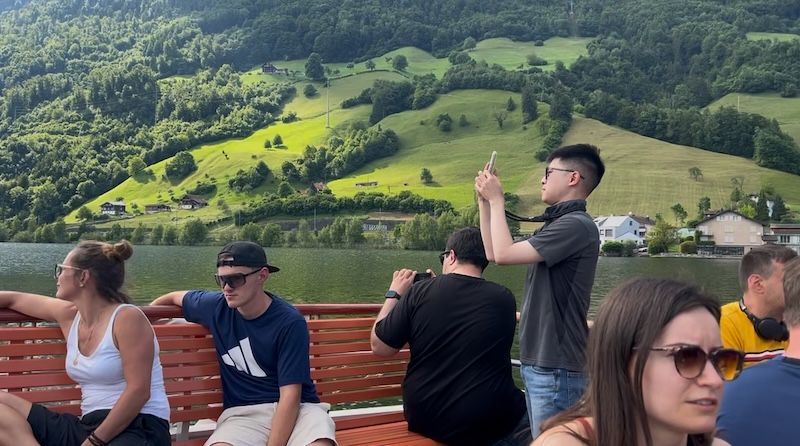 Passengers ride on a Lake Lucerne boat cruise with green mountains, lakeside homes, and forested hills in the background. The scenic view shows why Switzerland is worth visiting for easy day trips and lake cruises.