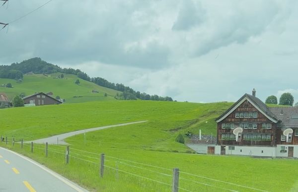 A roadside view of bright green Swiss countryside with rolling hills. farmhouses. fences. and cloudy skies. The peaceful scenery shows why Switzerland is worth visiting even between major stops.