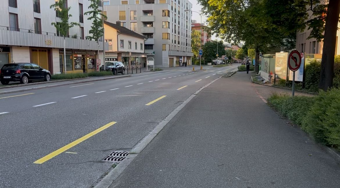 A quiet Swiss city street with a wide sidewalk, apartment buildings, parked cars, and trees in the evening light. The everyday street scene shows what it looks like to stay outside the main tourist areas when deciding is Switzerland worth visiting.