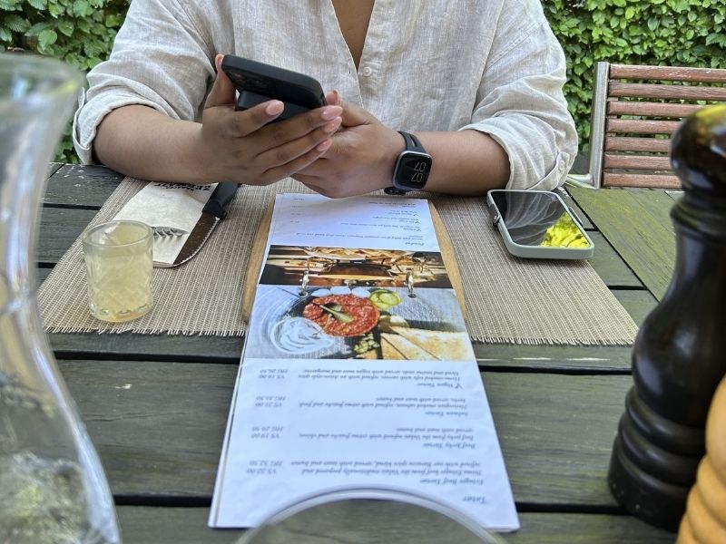 Jess sits at an outdoor restaurant table with her phone beside a printed menu. The menu and table setting help show what dinner costs can look like when deciding is Switzerland worth visiting.