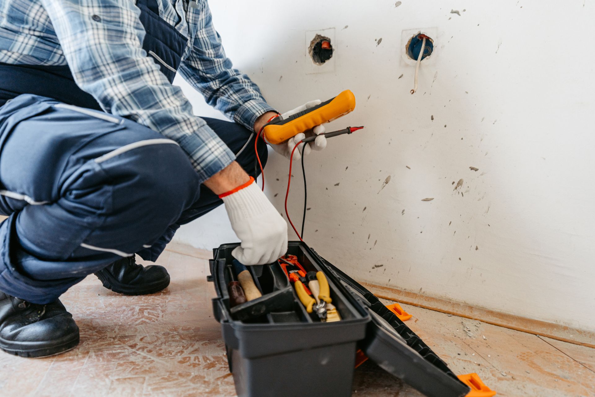 Un elettricista controlla il cablaggio con un multimetro, inginocchiato vicino alla cassetta degli attrezzi aperta e alle prese a muro.
