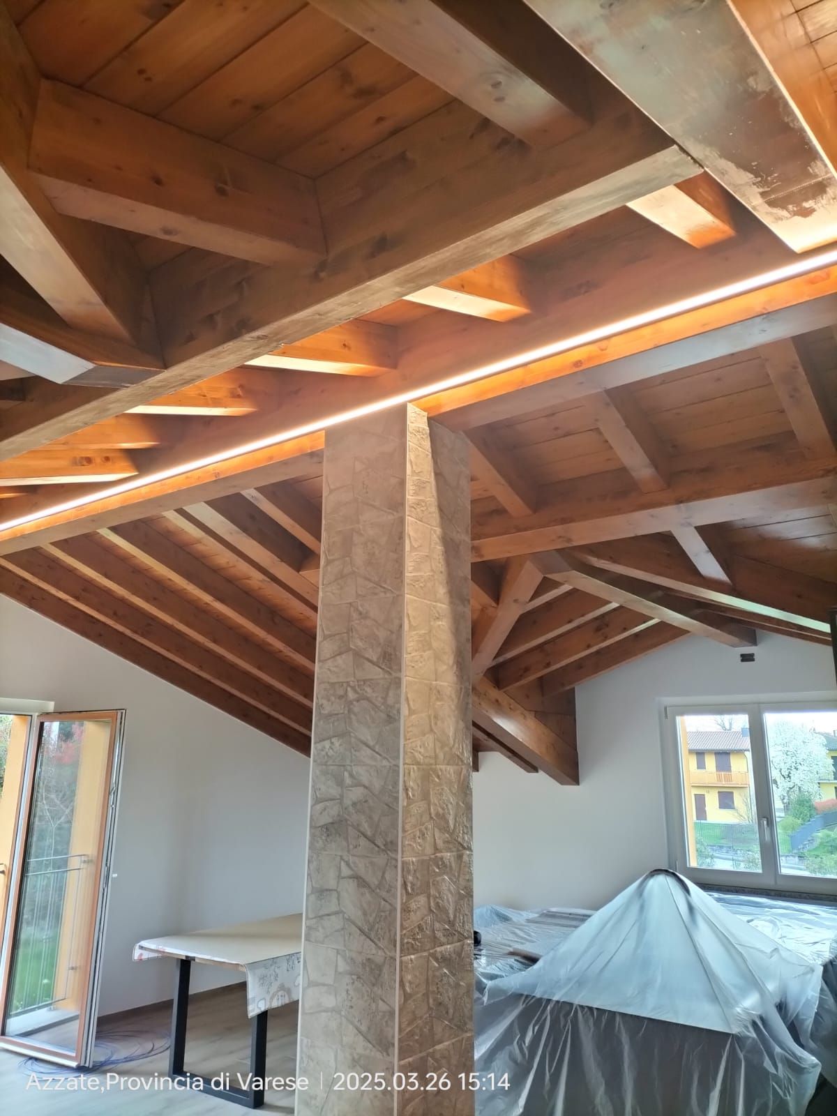 Soffitto con travi in legno e illuminazione a incasso sopra una colonna piastrellata. Interni luminosi con finestra e mobili rivestiti.