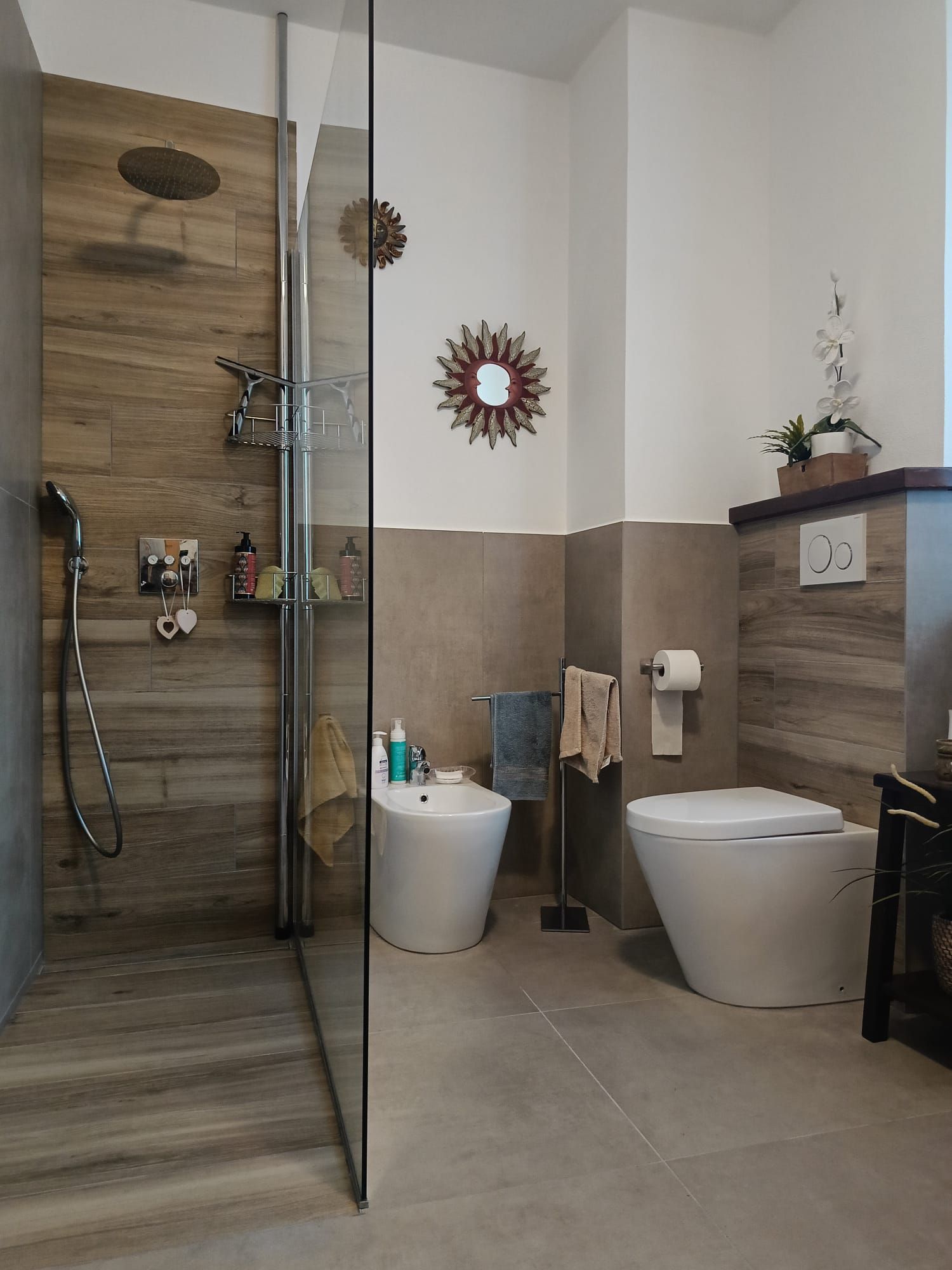 Bagno con doccia in vetro, WC, bidet e piastrelle effetto legno. Tonalità di grigio e marrone con luce solare.