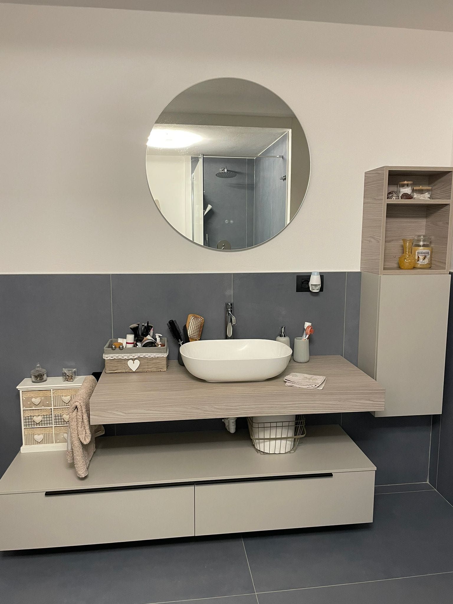 Bagno con uno specchio rotondo sopra un mobiletto sospeso con lavabo a appoggio e un mobiletto portaoggetti.