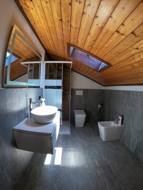 Bagno con soffitto in legno, lucernario e pareti e pavimento piastrellati grigi. Lavandino, WC e bidet bianchi.