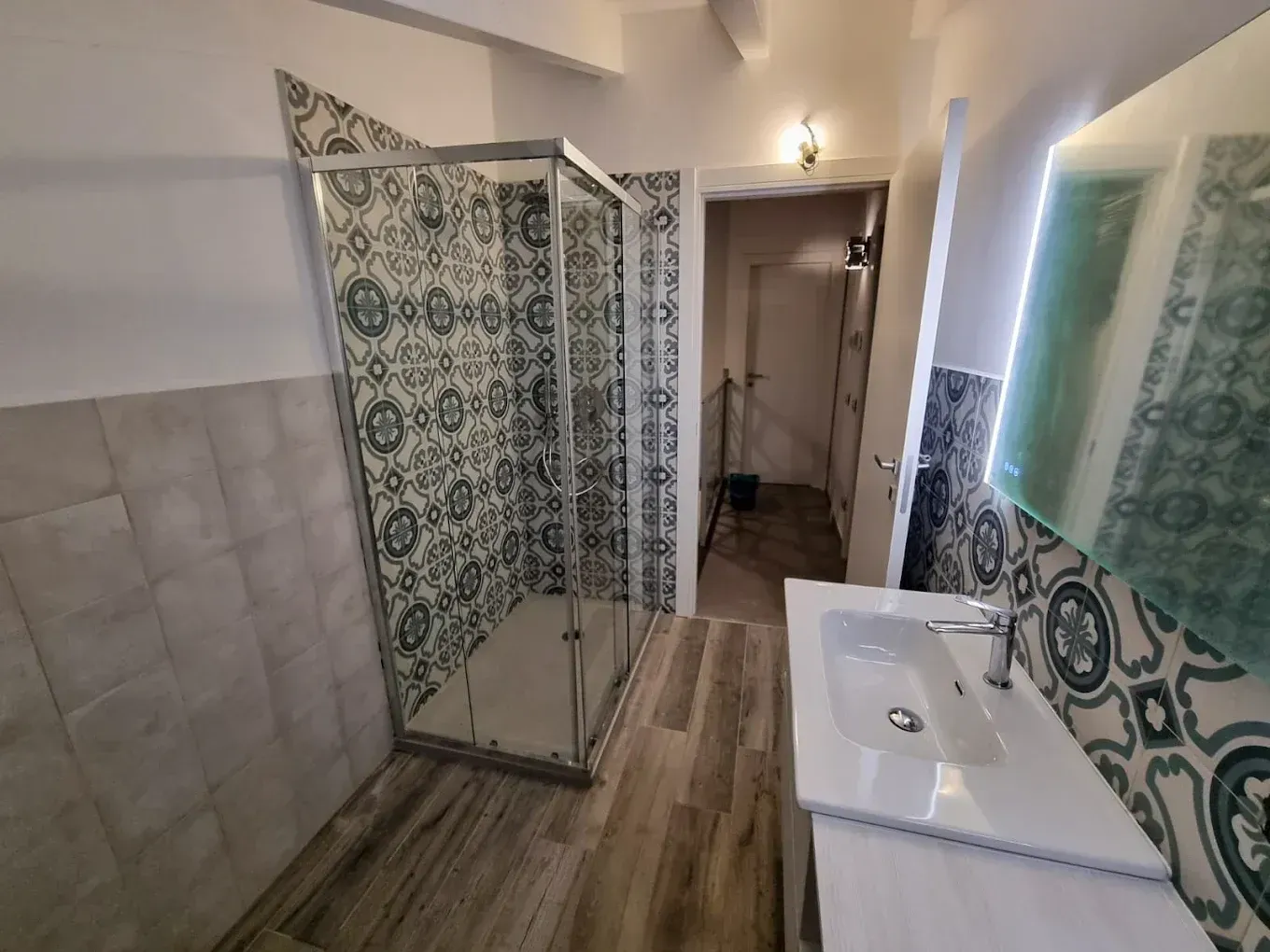 Bagno con doccia in vetro, piastrelle a motivi, mobile lavabo e porta che conduce a un'altra stanza.