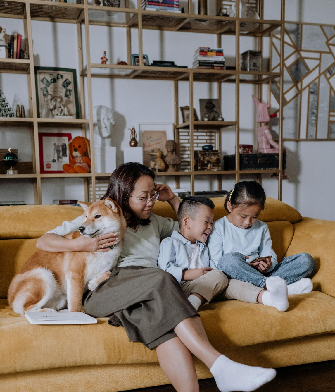 A woman and two children sit on a yellow sofa with a dog in a living room in AC cooled home.