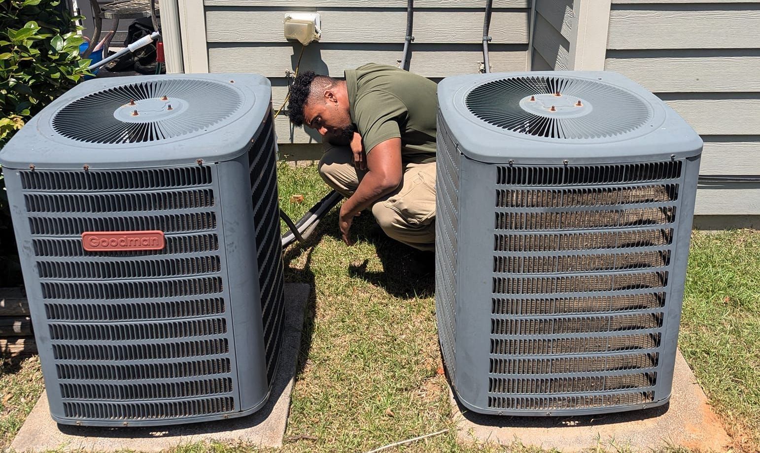 A man is kneeling down next to two air conditioners. Clayton, NC – Parker and Co Heating and Air, LLC