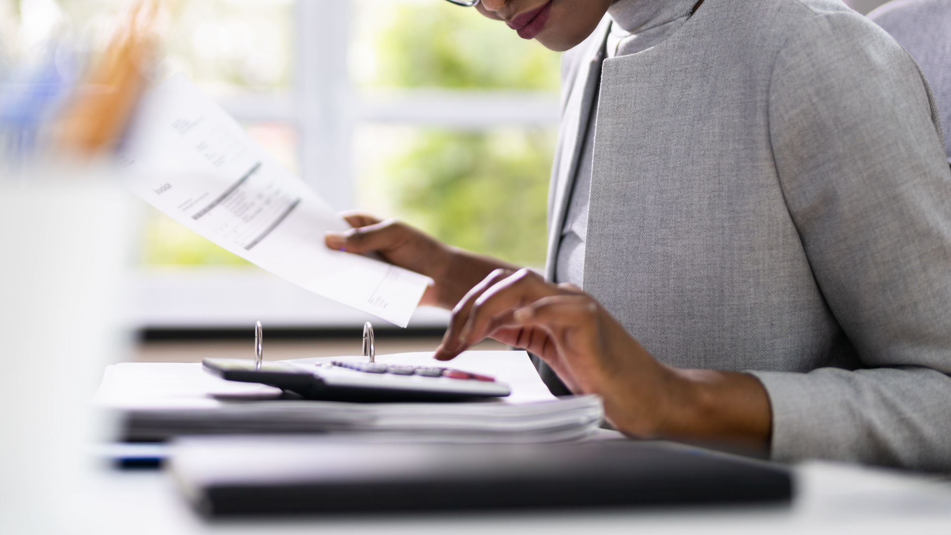 A woman is sitting at a desk using a calculator and a laptop.