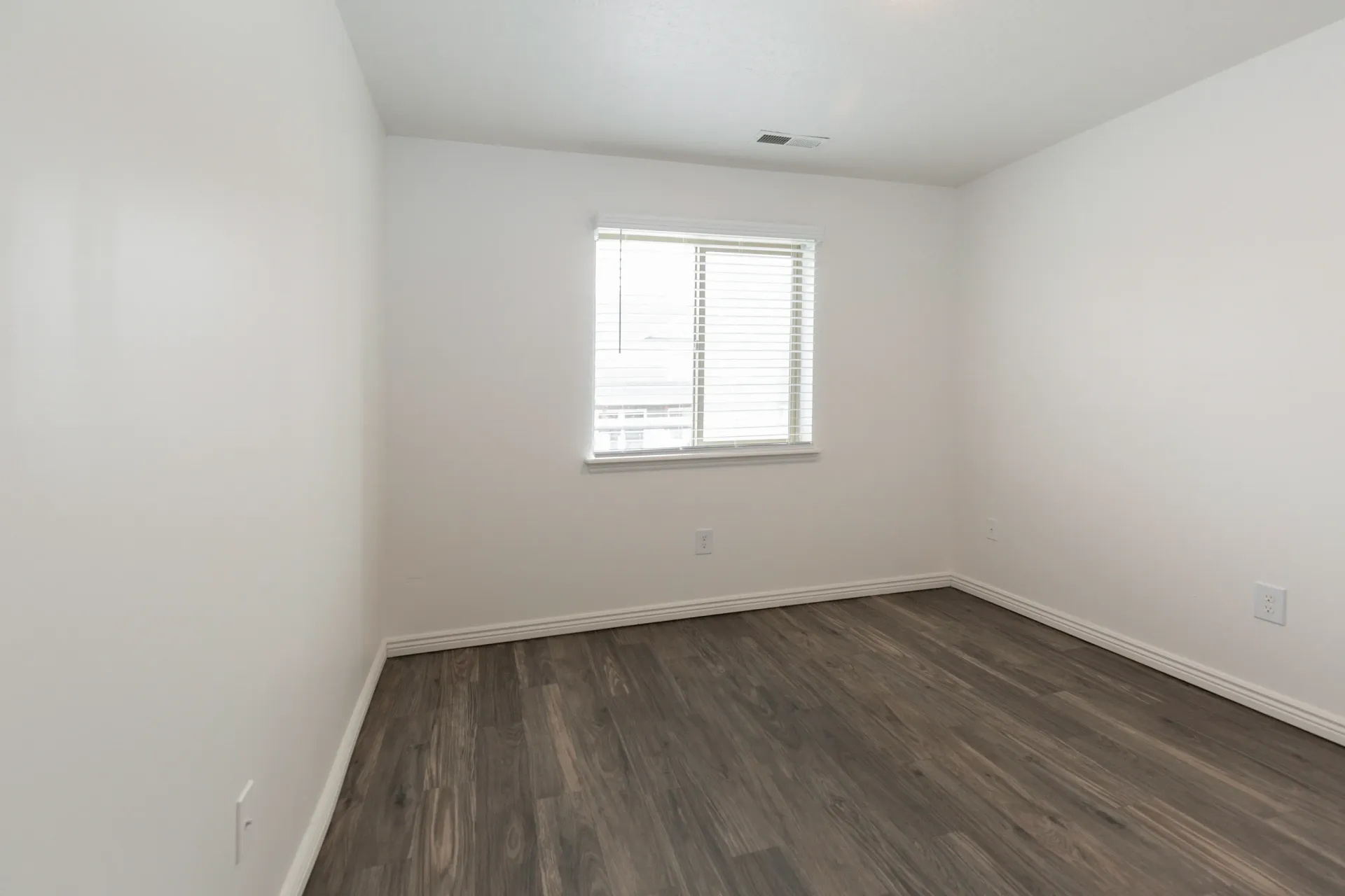 Empty room with a window and blinds, and dark wood floors.