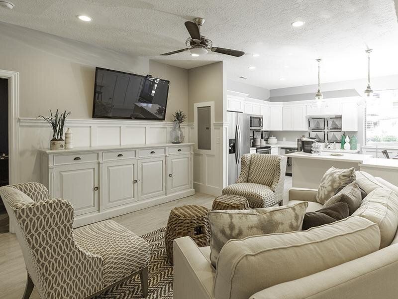 Bright living room with mounted TV, beige sofas, patterned chairs, and open view to a kitchen.