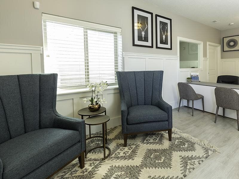 Bright living room with two blue chairs, patterned rug, side table, windows, and dining area beyond.