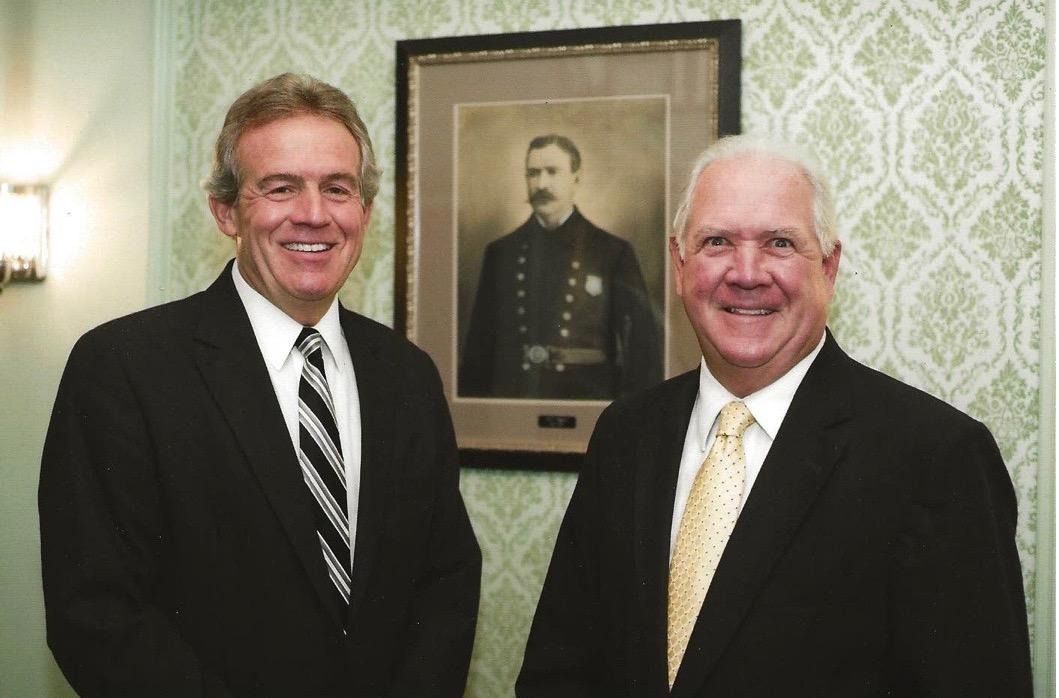 Two men in suits smile, standing by a framed portrait in a room with patterned wallpaper.
