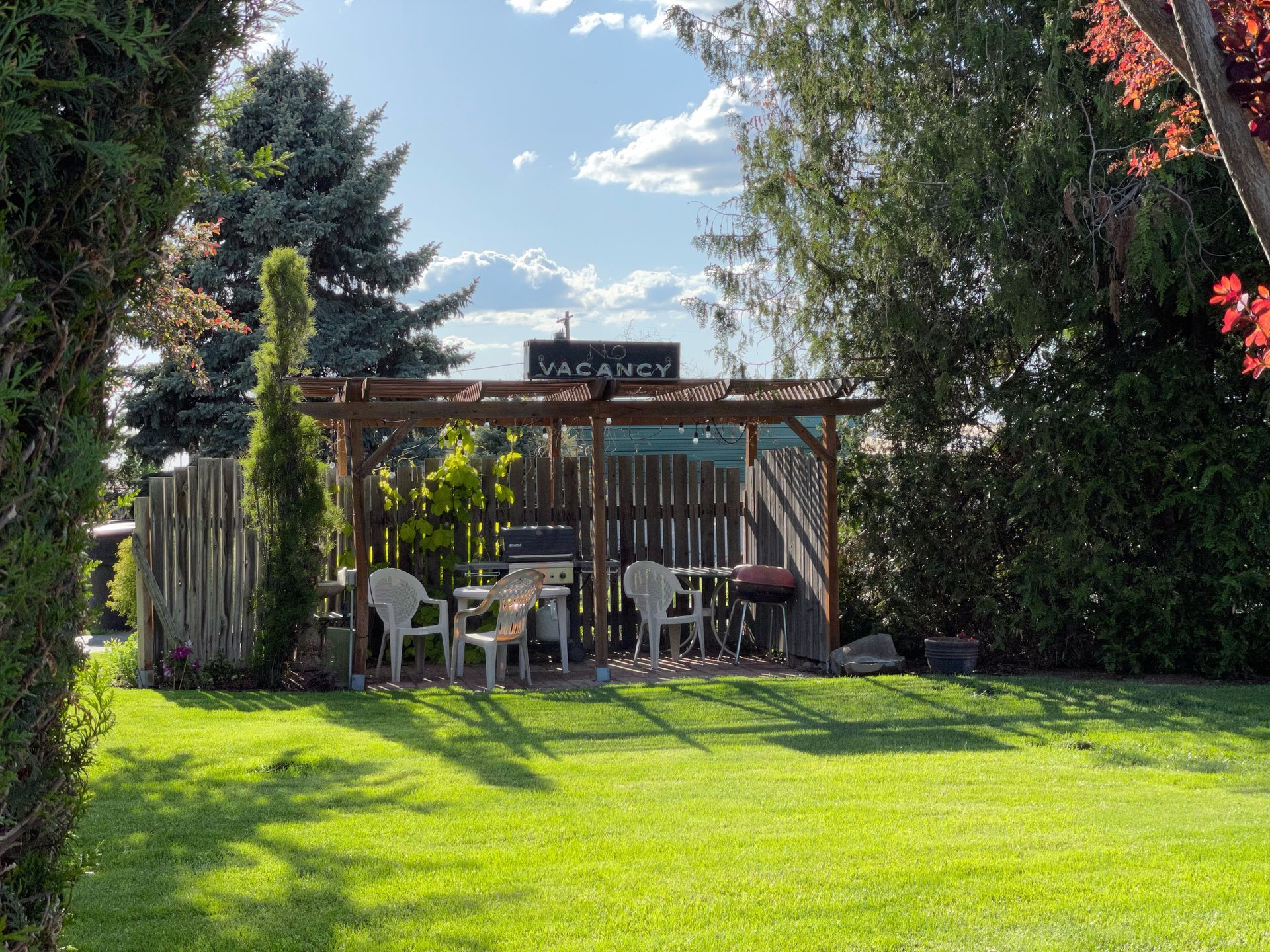 A lush green yard with a pergola , chairs , tables and a grill.