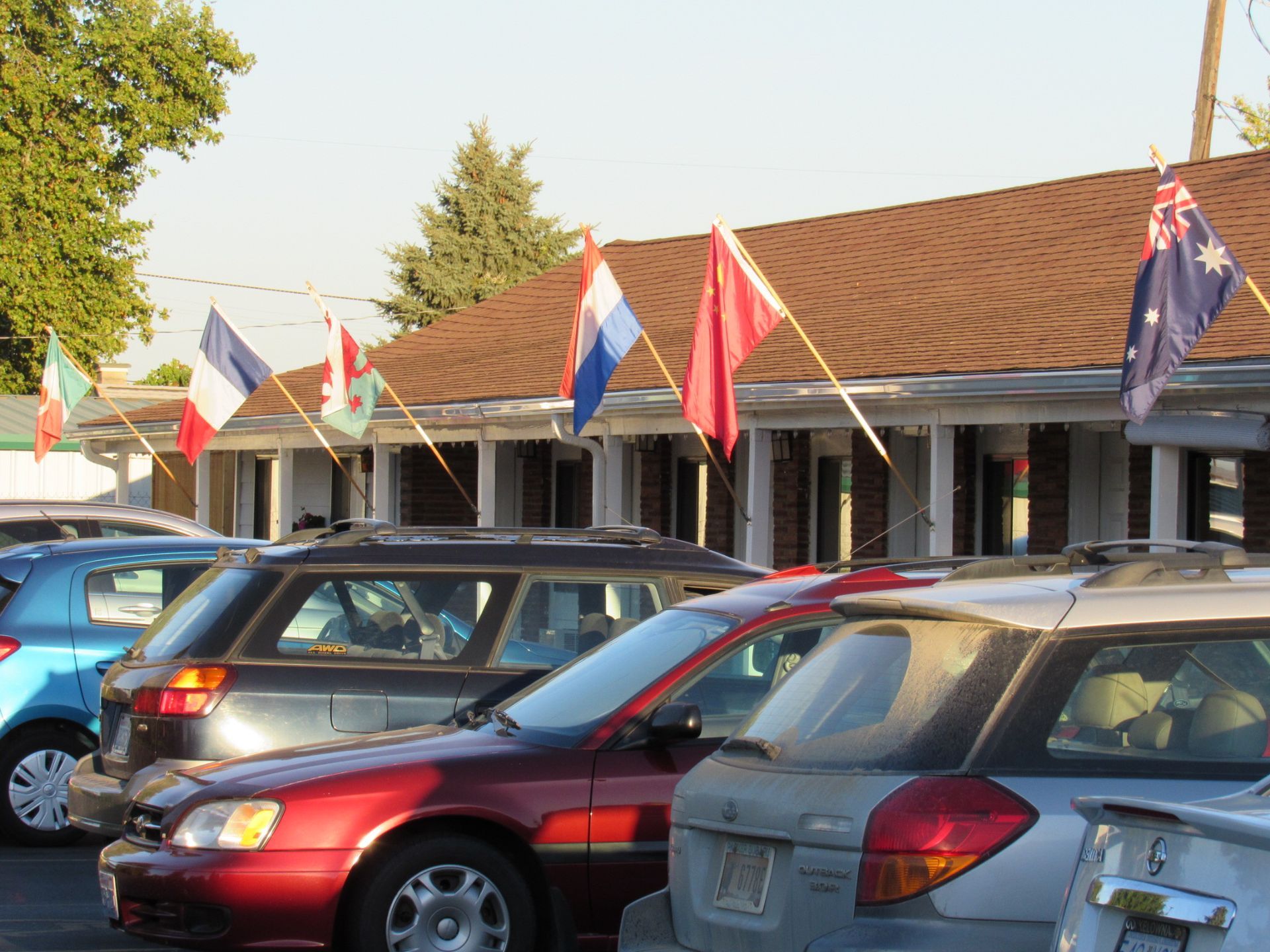 A row of cars are parked in front of a building with flags hanging from the roof
