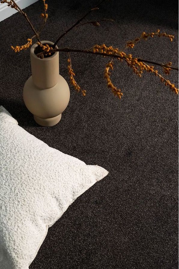 Beige vase with dried branches beside a white pillow on a dark textured carpet