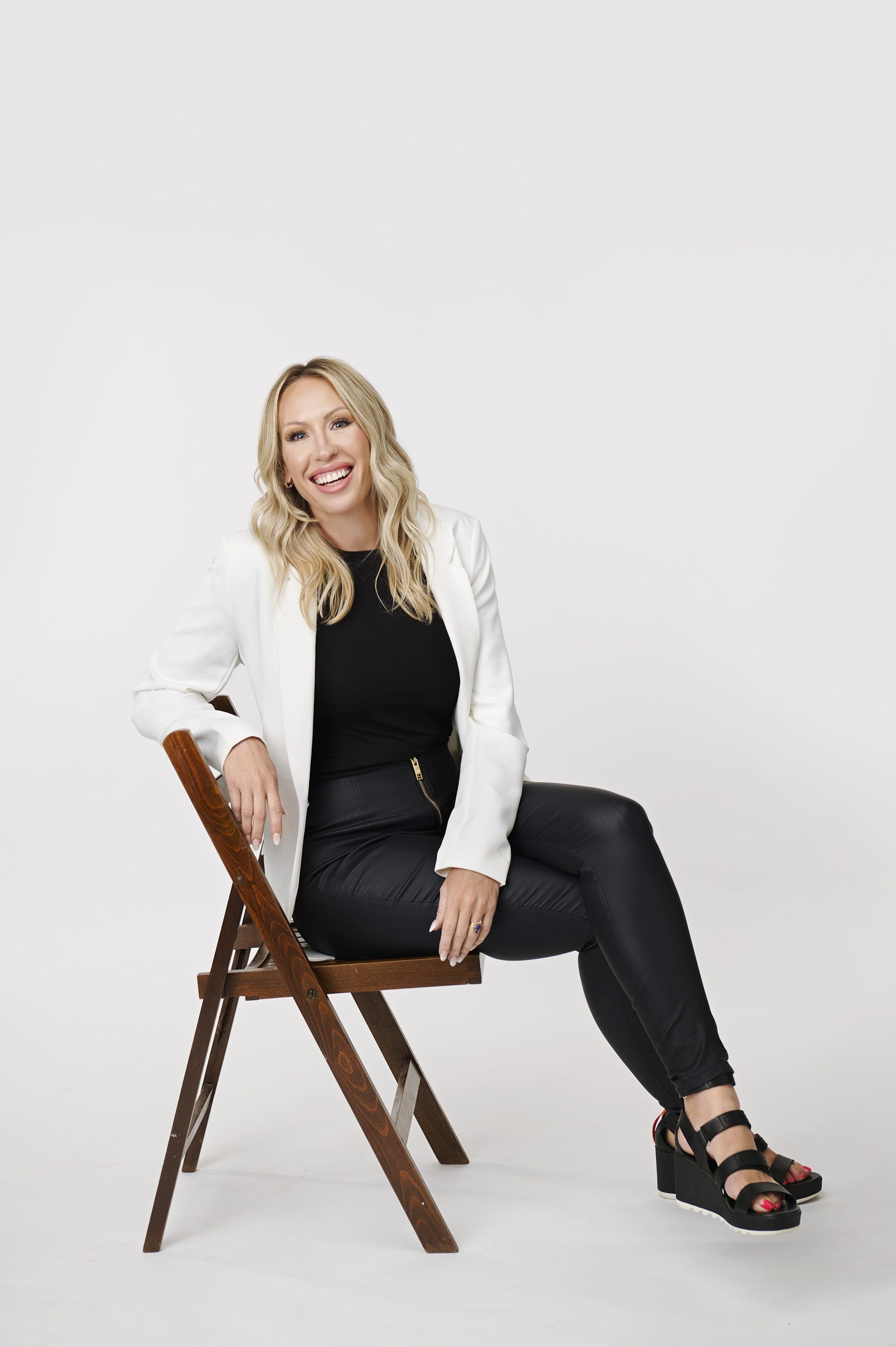 Blonde woman in white blazer sits on a chair, smiling. She wears black top, pants, and shoes. White background.