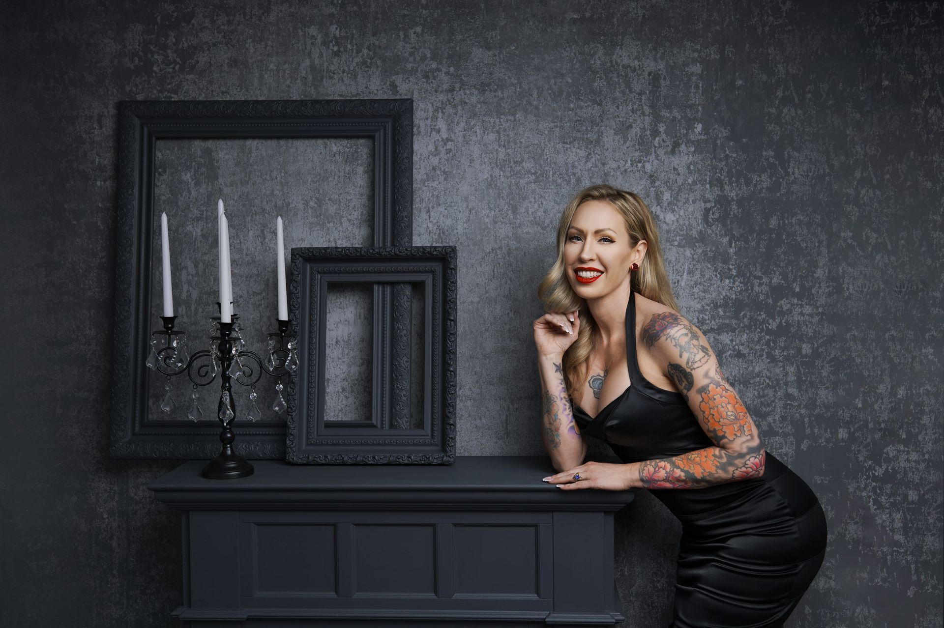 Woman with tattoos leans on a black mantle, smiles. Background: gray wall, candelabra, black frames.