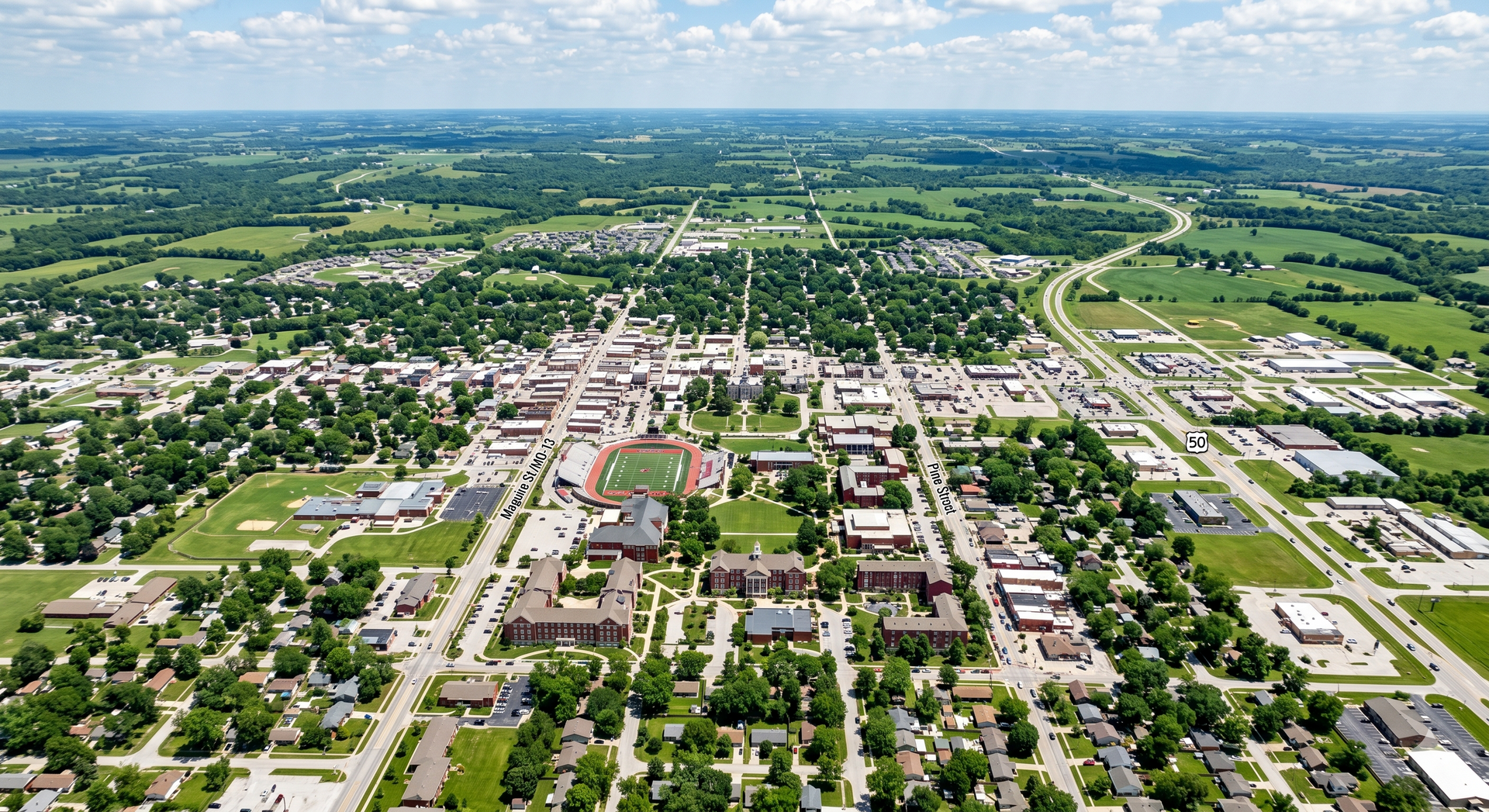 Aerial view of a suburban town with roads, houses, trees, and open fields under a blue sky