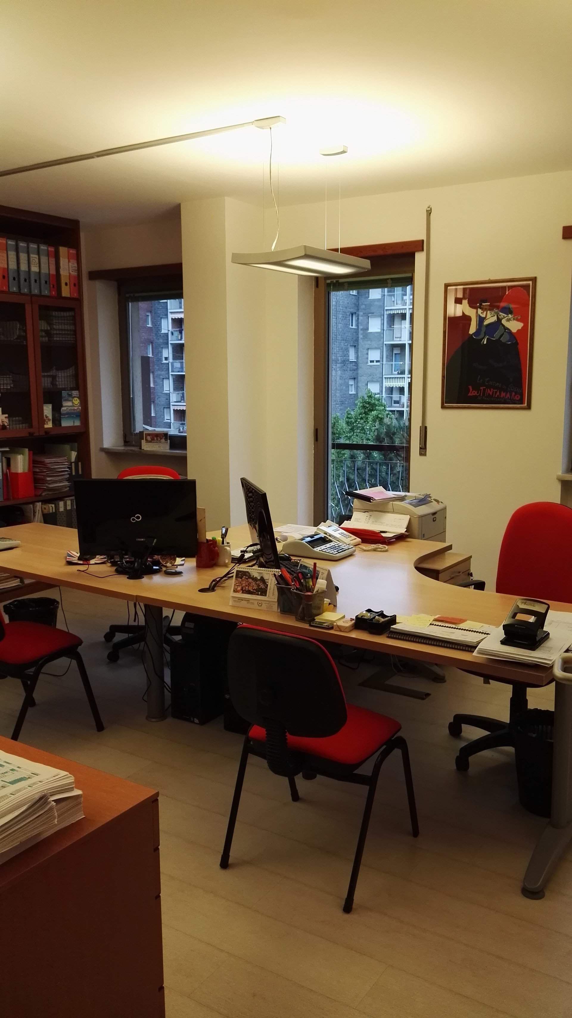 Un ufficio domestico disordinato con scrivanie, computer, sedie rosse e una finestra che si affaccia sul verde.