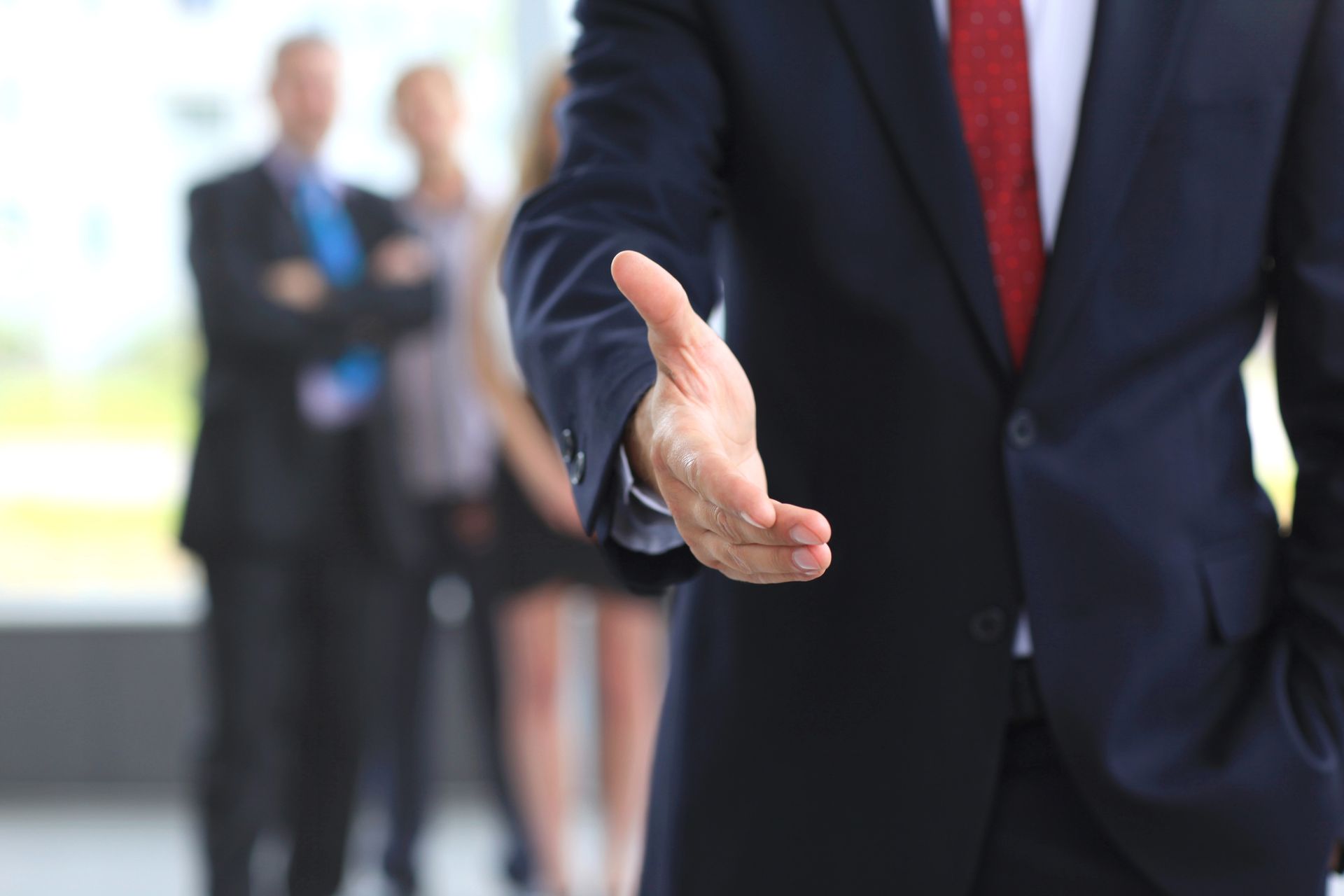 Uomo d'affari in abito scuro che porge la mano a qualcuno in ufficio, con i colleghi sfocati sullo sfondo.
