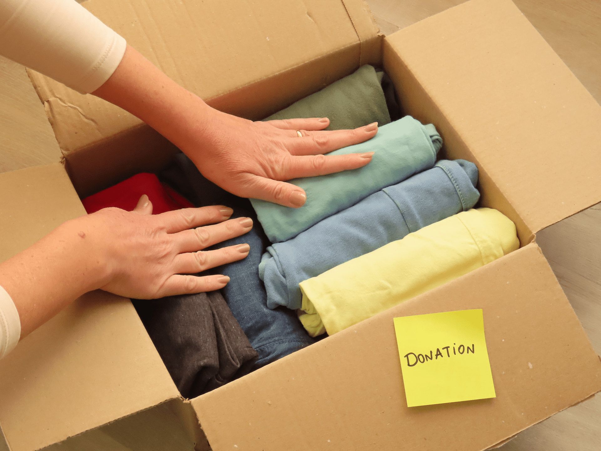 Hands arranging folded clothes in a cardboard donation box with a 