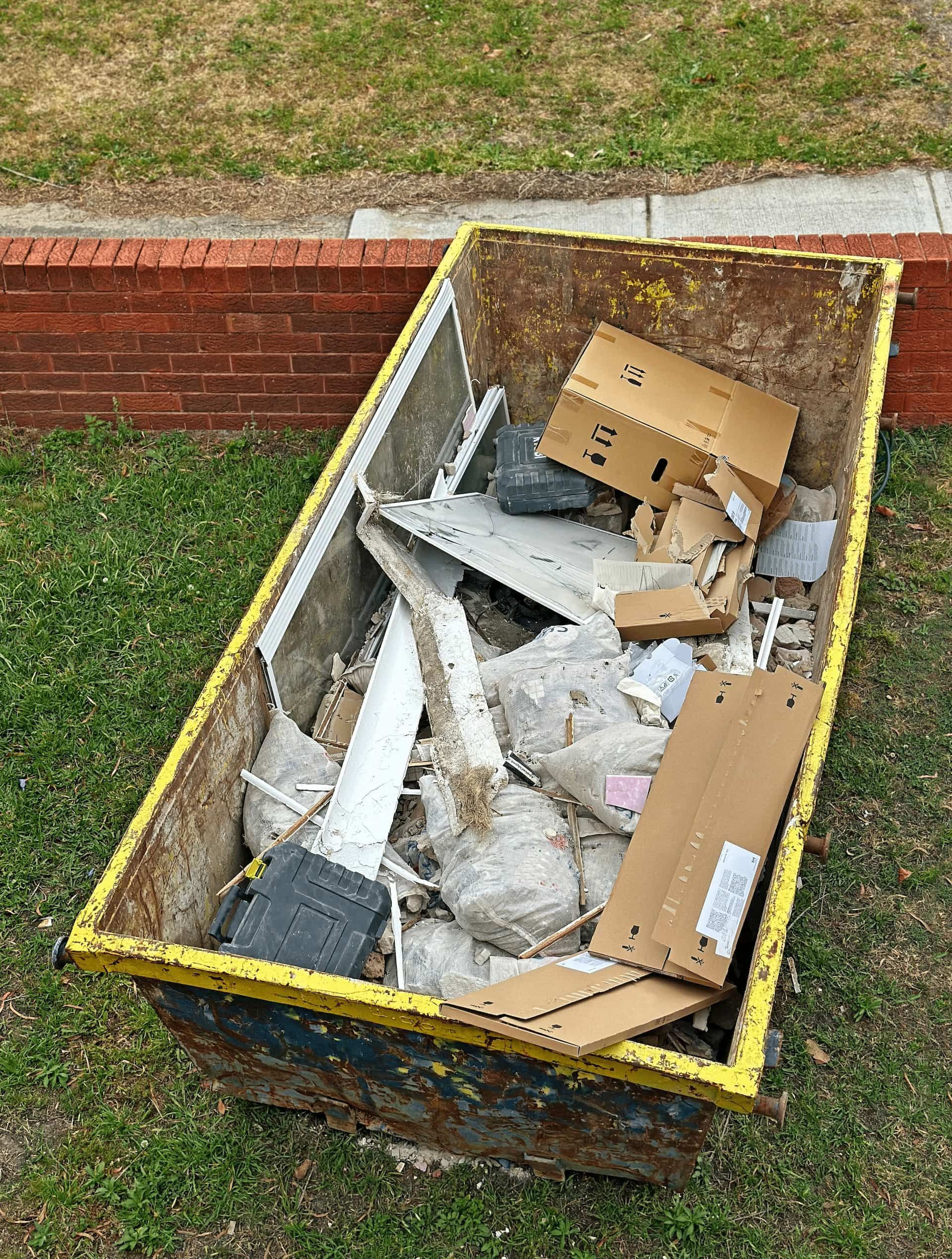 Rusty, yellow-rimmed dumpster filled with trash, including cardboard and metal, on green grass near brick wall.