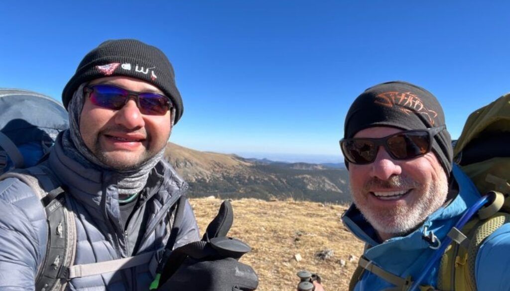 Two hikers smiling on a mountaintop, wearing sunglasses, beanies, and backpacks under a blue sky.