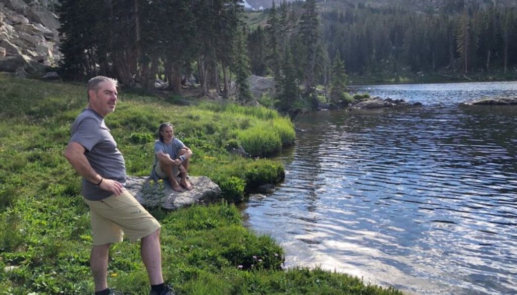 Man in shorts and tee looks at a lake, another person sits on a rock nearby, trees in the background.