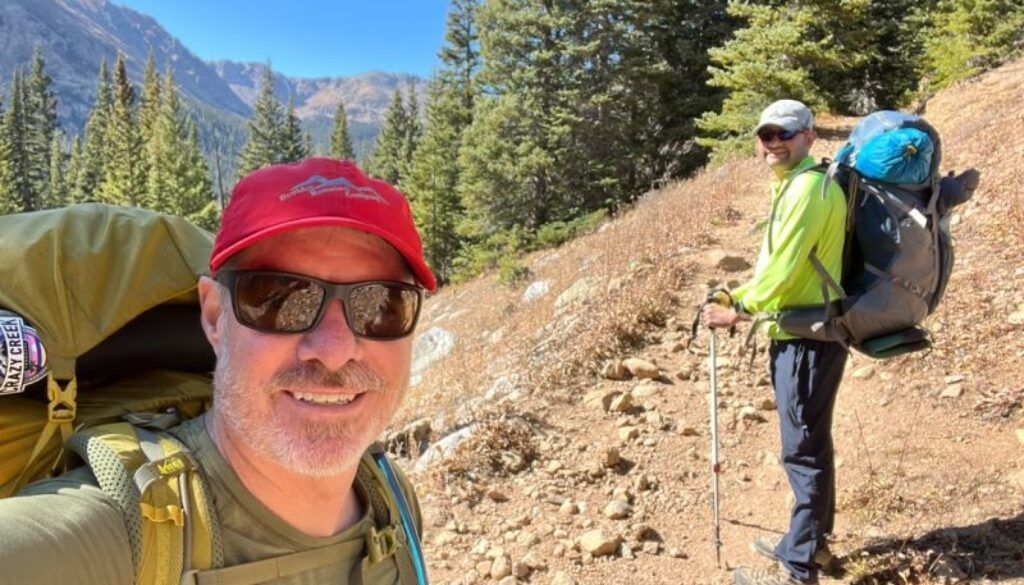 Two hikers on a mountain trail, one taking a selfie. Sunny day, green trees, backpacks.