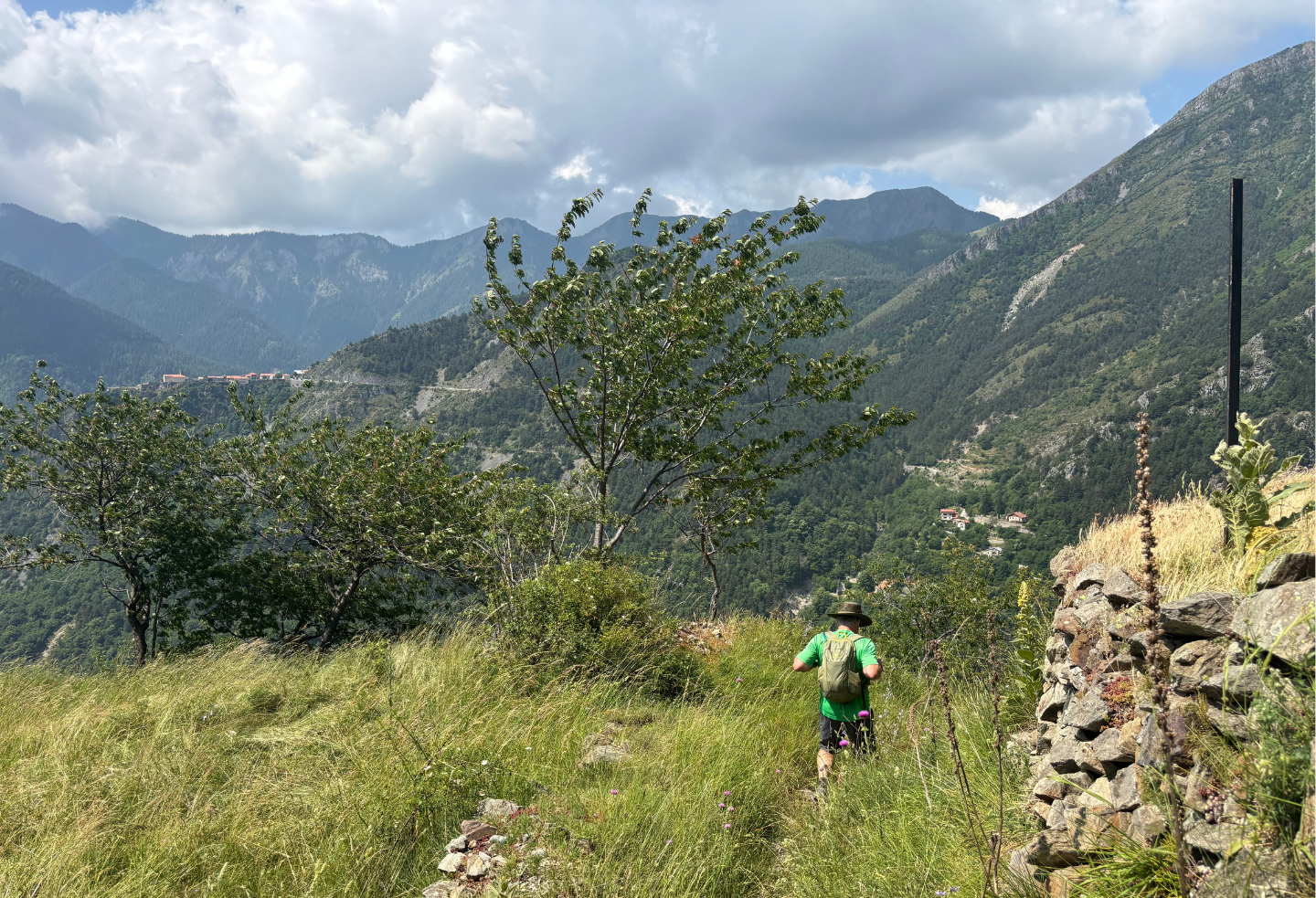 Person hiking on a grassy trail in mountainous landscape under a cloudy sky.